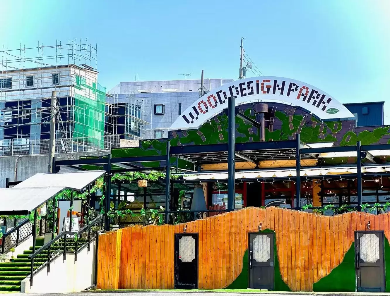 ウッドデザインパークという看板がある建物の外観写真。木製のフェンスがあり、緑の芝生が飾られている。階段と白い屋根が特徴的。