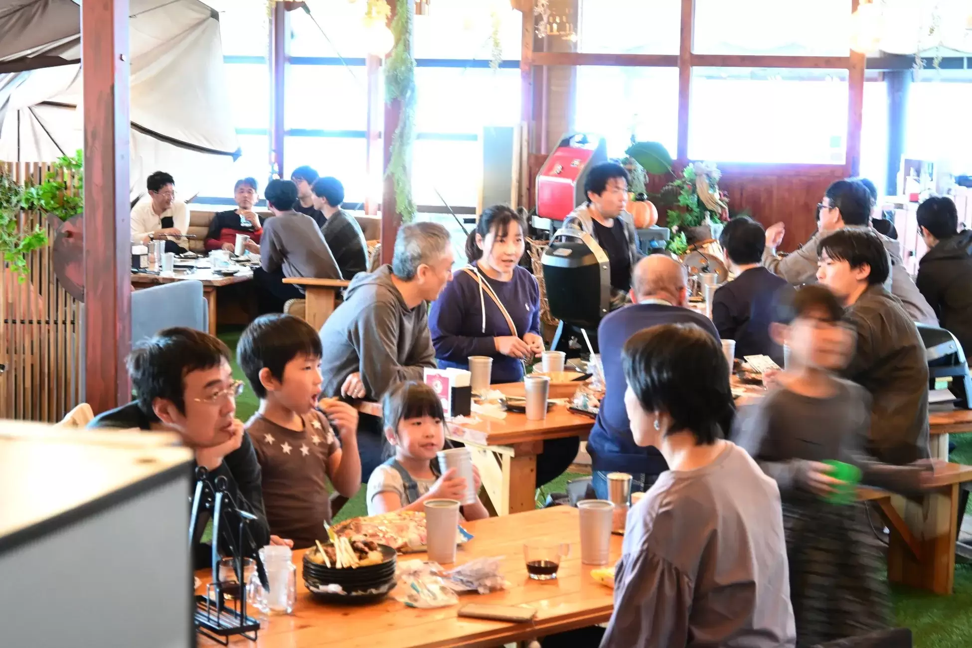 レストランで食事を楽しむ人々の様子を捉えた写真。木製のテーブルと椅子が配置され、人々は食事をしたり、会話を楽しんだりしている。背景には窓があり、自然光が差し込んでいる。