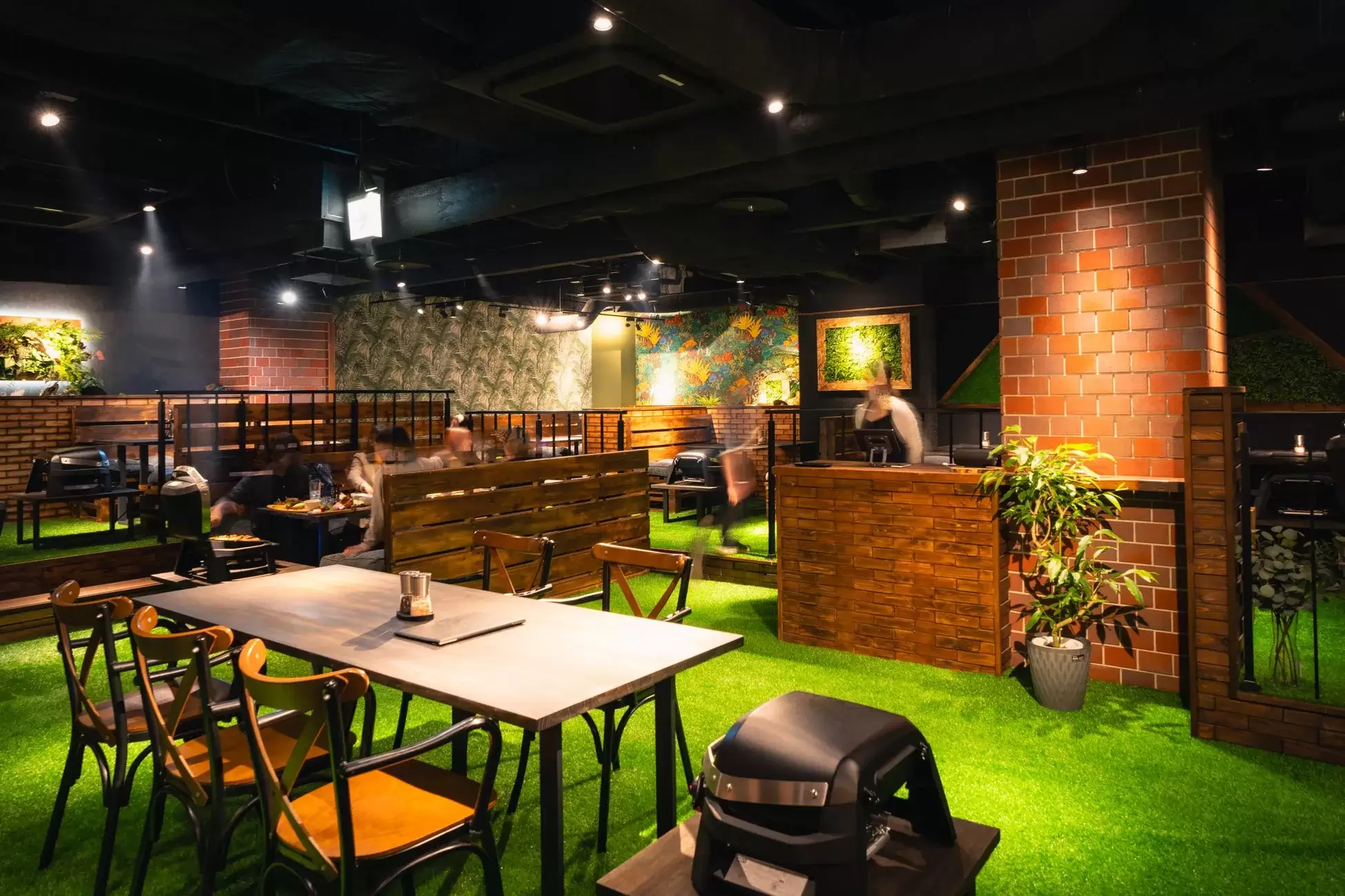 人工芝が敷かれたレストランの内部。レンガや木の壁、緑の装飾が施され、テーブルと椅子が配置されている。人々が食事を楽しんでいる様子が写っている。