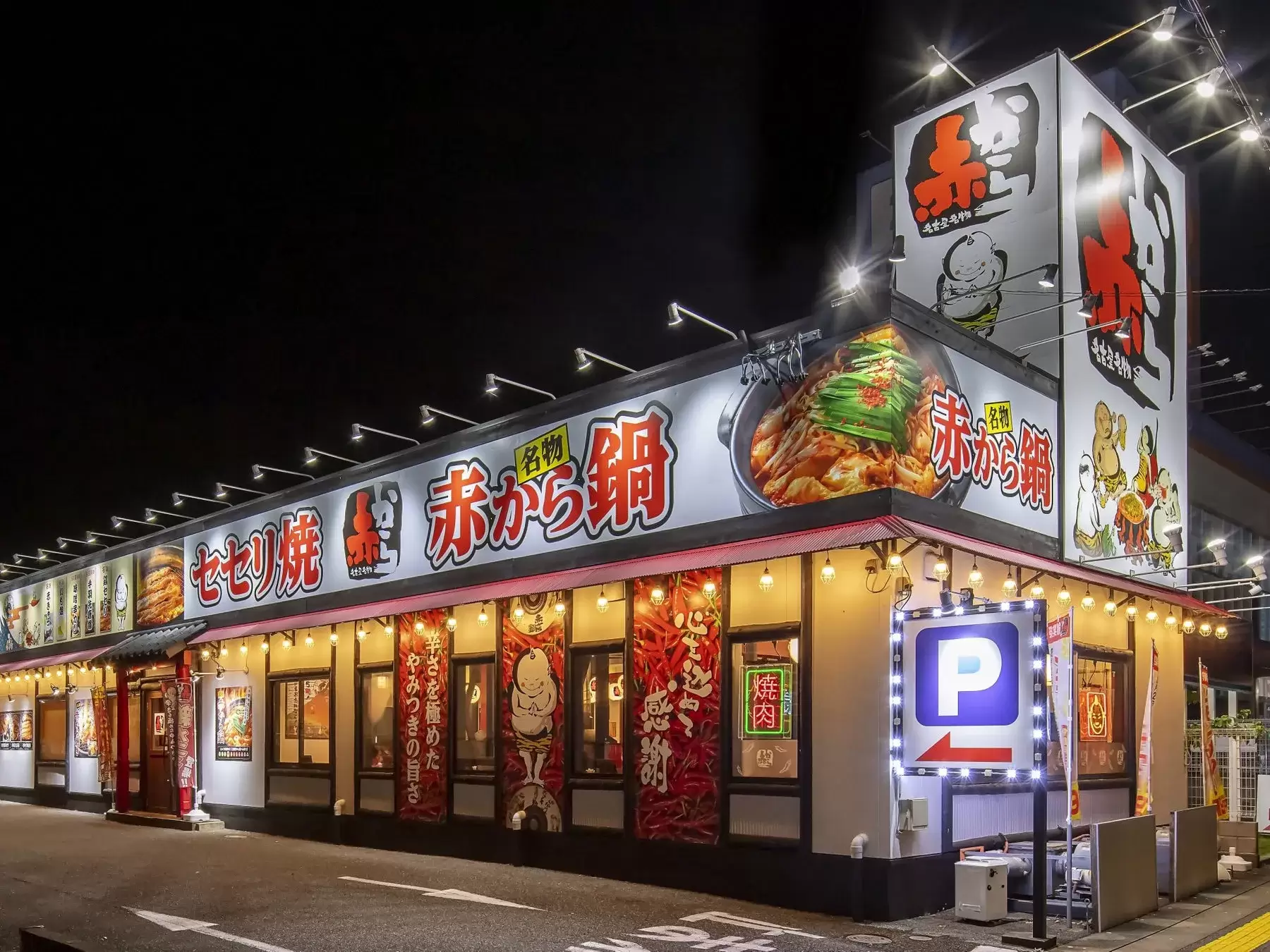 夜の街並みに佇む、赤と白を基調とした看板が目を引く飲食店。店名には「赤から鍋」と記され、鍋料理をメインに提供していることが伺える。駐車場案内の看板も設置されており、車での来店も可能。